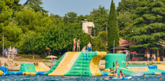 Kinder und Erwachsene planschen im Wasser und haben Spaß in dem See. Es ist Sommer, die Temperaturen sind hoch, es ist heiß. Auf dem Wasser befinden sich eine große aufblasbare Hüpfburg und ein Spielplatz.