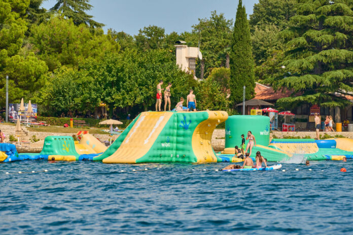 Kinder und Erwachsene planschen im Wasser und haben Spaß in dem See. Es ist Sommer, die Temperaturen sind hoch, es ist heiß. Auf dem Wasser befinden sich eine große aufblasbare Hüpfburg und ein Spielplatz.