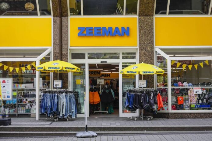 Eine Filiale der niederländischen Einzelhandelskette "Zemann" im typischen gelb-blauen Design. Vor dem Geschäft hängen Jeans und andere Hosen unter Schirmen. Preisschilder locken mit Angeboten.