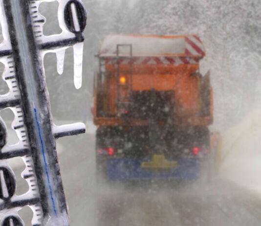 Blick auf eine vereiste Straße mit einem Lkw, der sich seinen Weg durch einen Schneesturm bahnt. Davor erkennt man ein Thermometer, welches ebenfalls mit Eis bedeckt ist. Es ist offenbar tiefster Winter und sehr kalt.