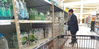 Der Blick in einen Supermarkt. In der Abteilung für abgepacktes Mineralwasser. Verschiedene bekannte Wassermarken stehen im Regal, in Plastikflaschen abgefüllt. Eine Frau greift gerade nach einem Sechserpack Wasser und will dieses offenbar in ihren Einkaufswagen legen.