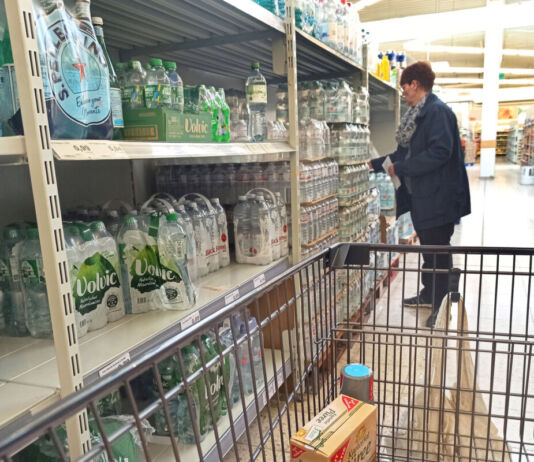 Der Blick in einen Supermarkt. In der Abteilung für abgepacktes Mineralwasser. Verschiedene bekannte Wassermarken stehen im Regal, in Plastikflaschen abgefüllt. Eine Frau greift gerade nach einem Sechserpack Wasser und will dieses offenbar in ihren Einkaufswagen legen.