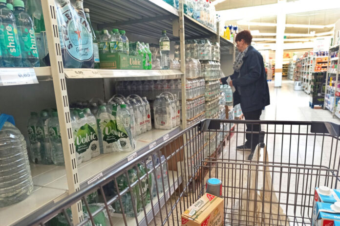 Der Blick in einen Supermarkt. In der Abteilung für abgepacktes Mineralwasser. Verschiedene bekannte Wassermarken stehen im Regal, in Plastikflaschen abgefüllt. Eine Frau greift gerade nach einem Sechserpack Wasser und will dieses offenbar in ihren Einkaufswagen legen.