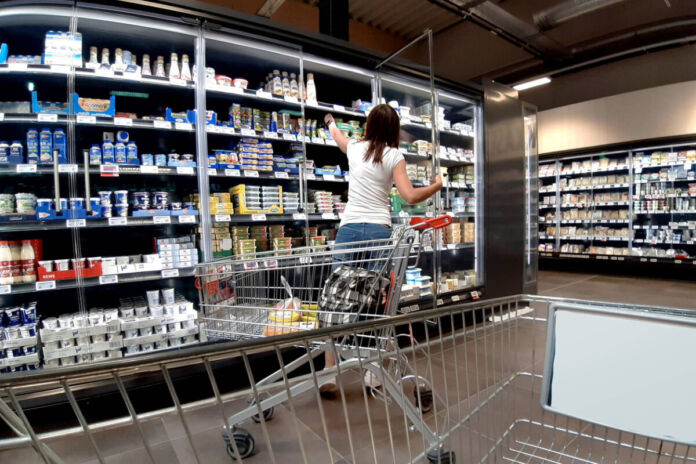 Ein Blick in einen Supermarkt oder einen Discounter. Eine Frau steht vor einem Regal mit Milchprodukten und greift nach einem der oberen Produkte. Vor ihr steht ein Einkaufswagen mit einigen Lebensmitteln. Die Regale sehen gut gefüllt aus.