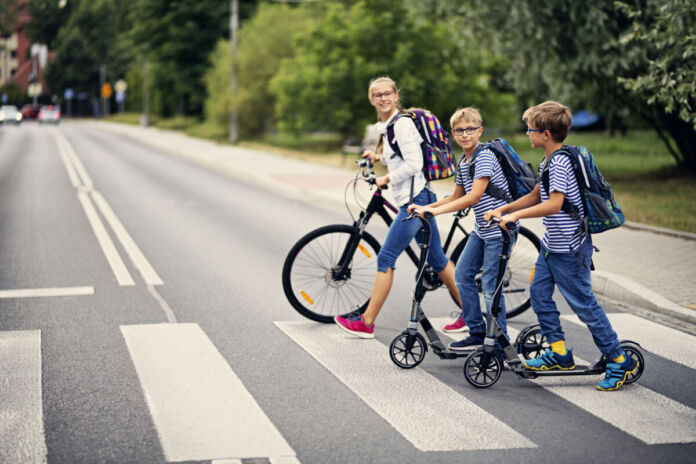 Drei Kinder überqueren einen Zebrastreifen. Sie sind wohl auf dem Weg zur Schule. Zwei Jungs fahren Roller, ein Mädchen fährt Fahrrad. Auf der Straße müssen sie auf Autos aufpassen.