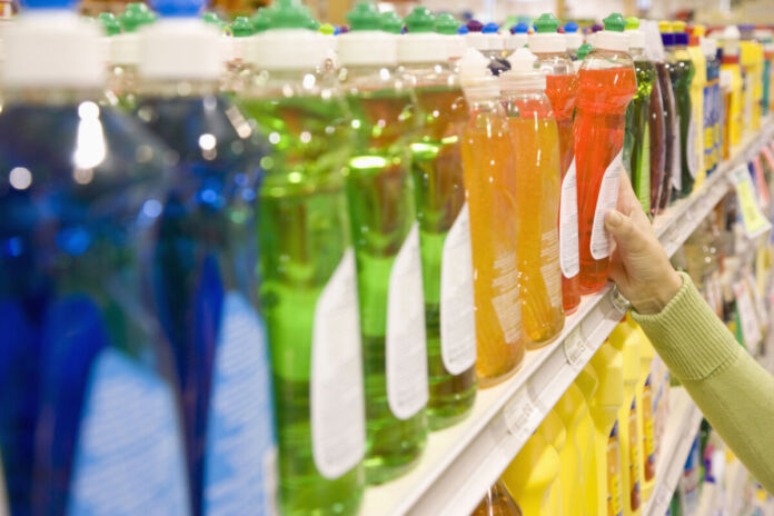 Viele verschiedene, bunte Geschirrspülmittel in Plastikverpackungen in einem Supermarktregal. Eine Hand greift zu einem der Geschirrspülmittel; der Kunde hat sich entschieden und möchte das Geschirrspülmittel kaufen.