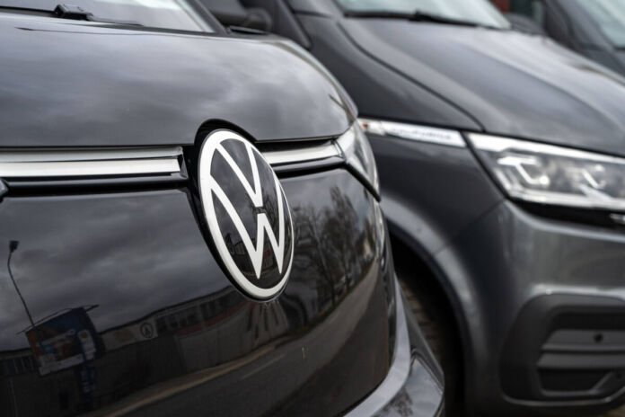 Ein schwarzes Elektroauto von VW steht auf einem Parkplatz in einer Reihe von anderen Fahrzeugen. Der Wagen ist ein VW ID Buzz. Die Karosserie glänzt und das Logo befindet sich an der Frontseite des E-Autos.