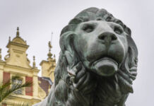 Ein aus Bronze gegossener Löwe. Die Skulptur ist aus massivem Metall kunstvoll gefertigt und ziert den Eingang vor einem Schloss, das man im Hintergrund erkennt. Er stammt aus einer Kunstgießerei.