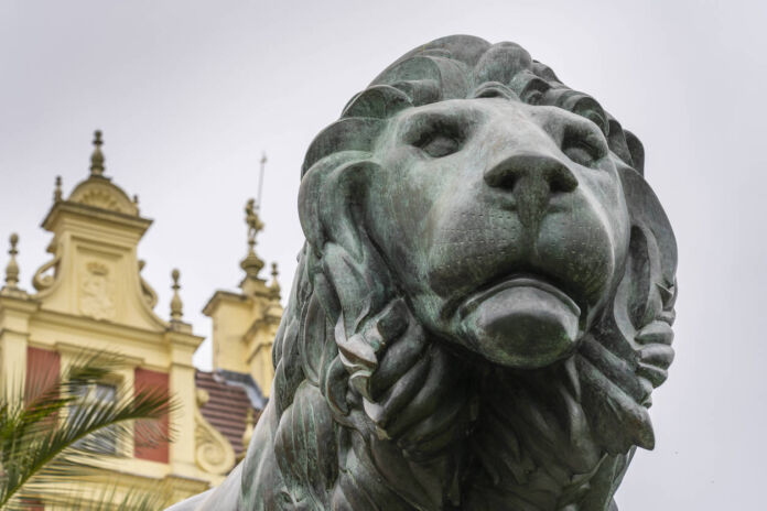 Ein aus Bronze gegossener Löwe. Die Skulptur ist aus massivem Metall kunstvoll gefertigt und ziert den Eingang vor einem Schloss, das man im Hintergrund erkennt. Er stammt aus einer Kunstgießerei.